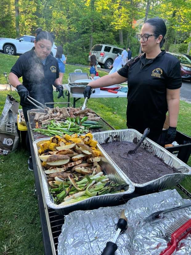 Two cooks working the grill with trays of corn, potatoes and beans